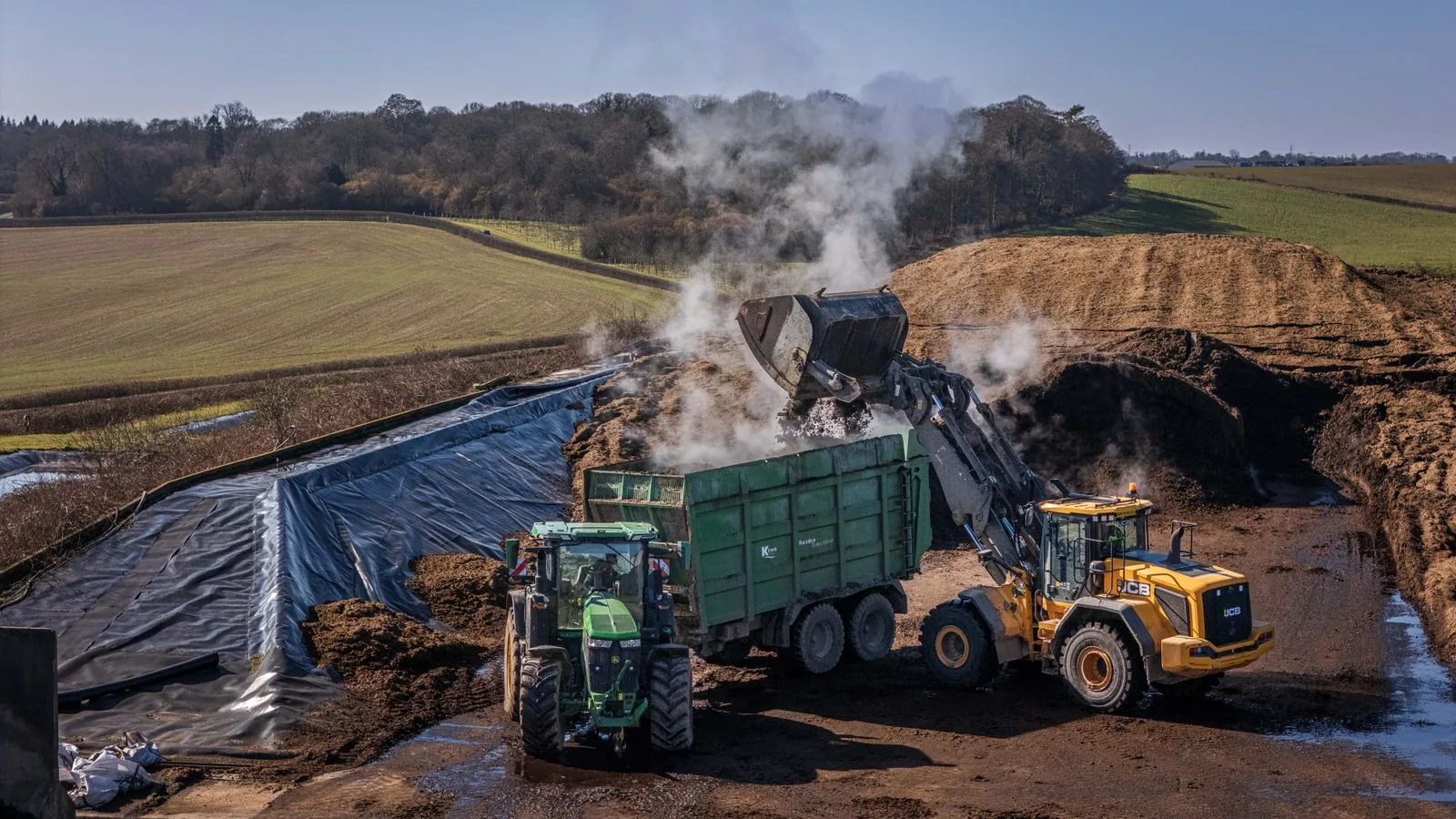 A front loader turning a long, steaming windrow of compost at an outdoor composting facility.